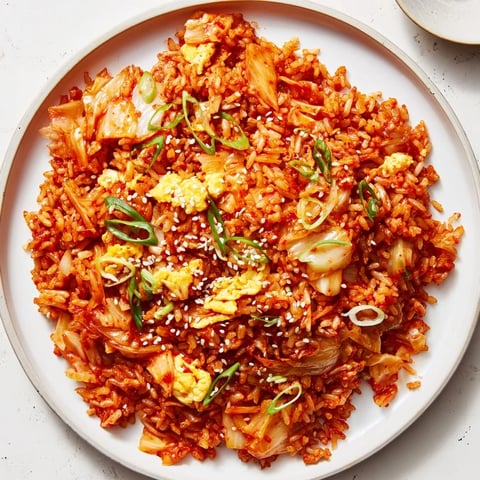 A close-up of sizzling Spicy Kimchi Fried Rice in a skillet, with vibrant orange kimchi pieces and scrambled egg ribbons mixing through the hot rice.