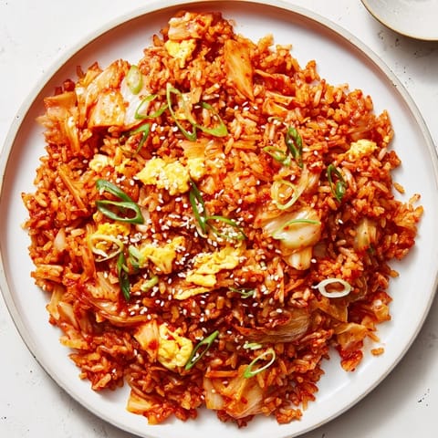 A close-up of sizzling Spicy Kimchi Fried Rice in a skillet, with vibrant orange kimchi pieces and scrambled egg ribbons mixing through the hot rice.
