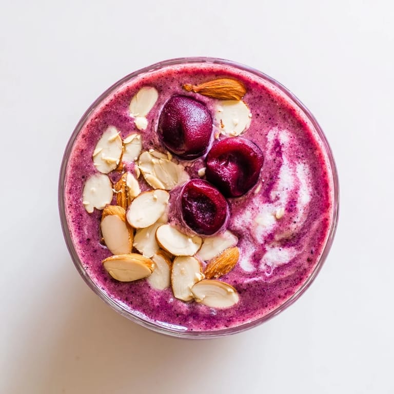 Two tall glasses of Cherry Almond Smoothie with a cherry garnish, set on a marble countertop alongside sliced bananas and measuring cups.